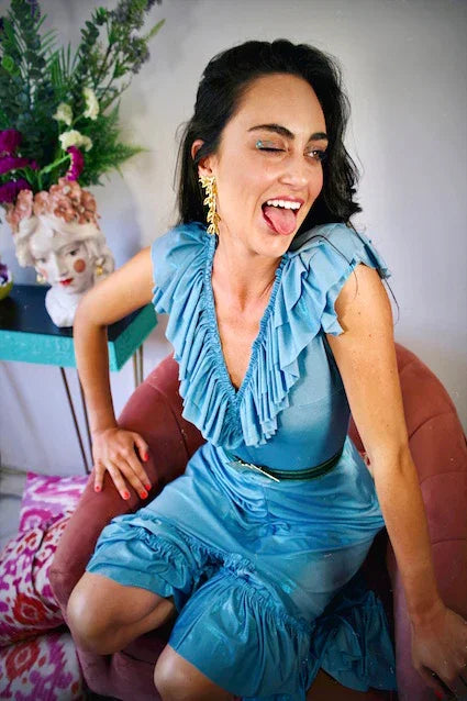 Mujer sentada en sillón rosa, con vestido azul celeste y maquillaje brillante, sonriente.