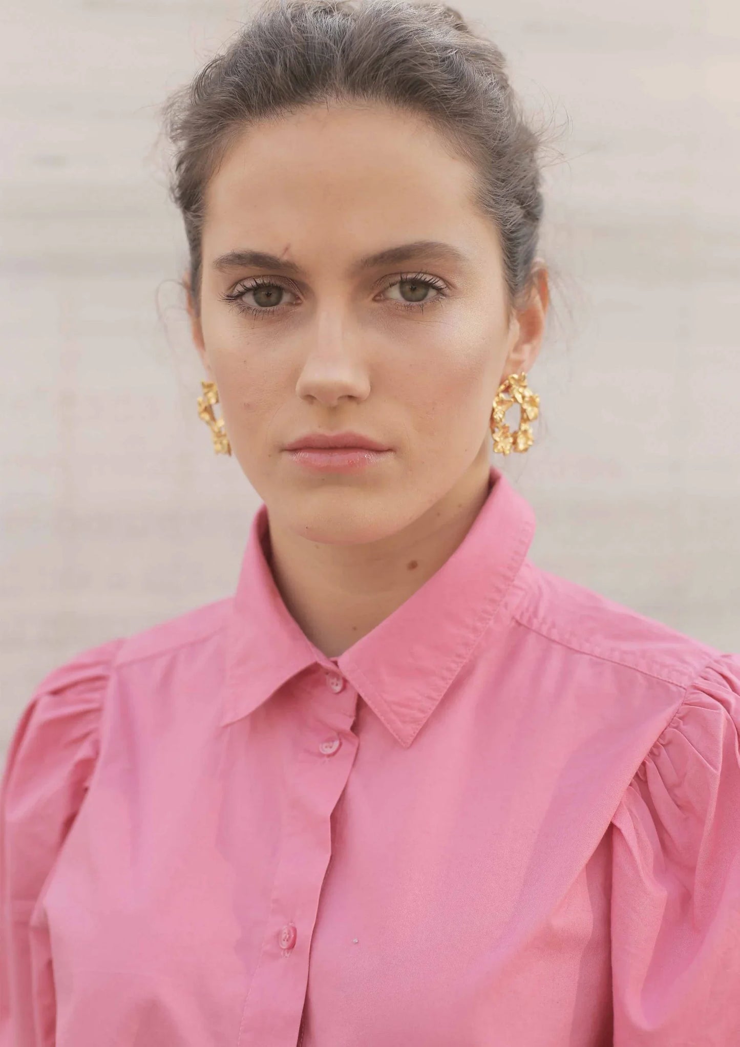 Mujer con camisa rosa y pendientes dorados grandes, fondo claro, moda femenina elegante.