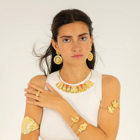 Mujer luciendo collar dorado con detalles rosas, pendientes y brazaletes dorados, fondo neutro