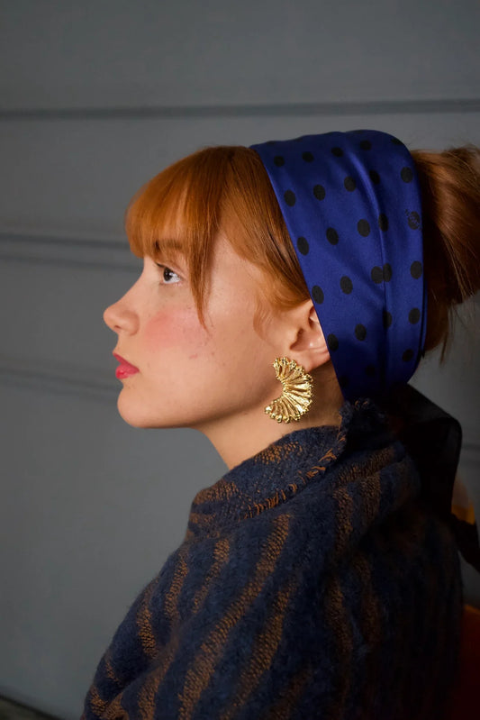 Mujer con bandana azul de lunares y pendientes dorados, fondo gris elegante.