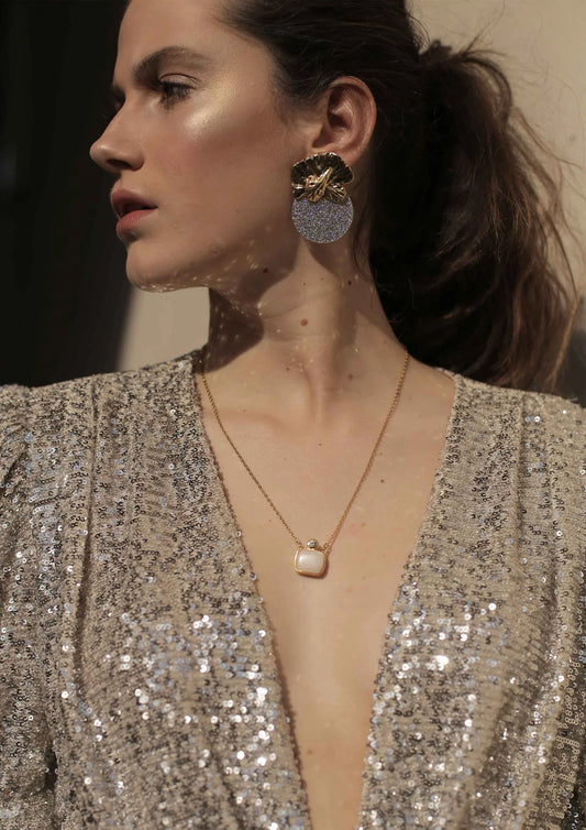Mujer con pendientes dorados grandes de orquídea y collar colgante sobre vestido brillante.