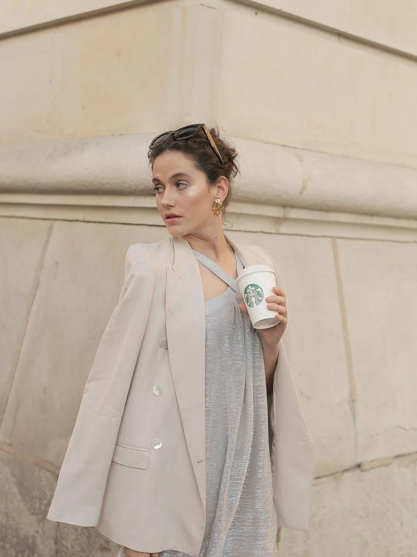 Mujer elegante con blazer beige y vestido plateado sosteniendo café en ambiente urbano